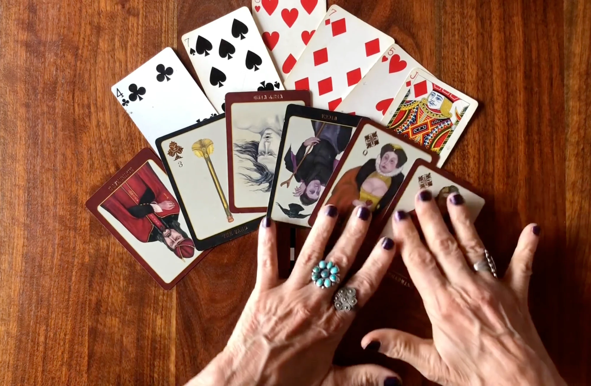 Ana's hands resting on a spread of playing cards and oracle cards on a wooden table